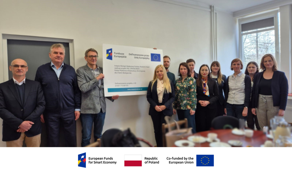 A group of people are in a room. The men are holding a plaque with information about the POL-OPENSCREEN project funding. They are all smiling.