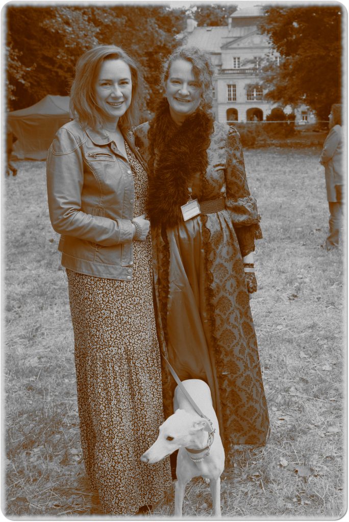 Sepia photo: two smiling women and a dog; behind them, a meadow and a palace