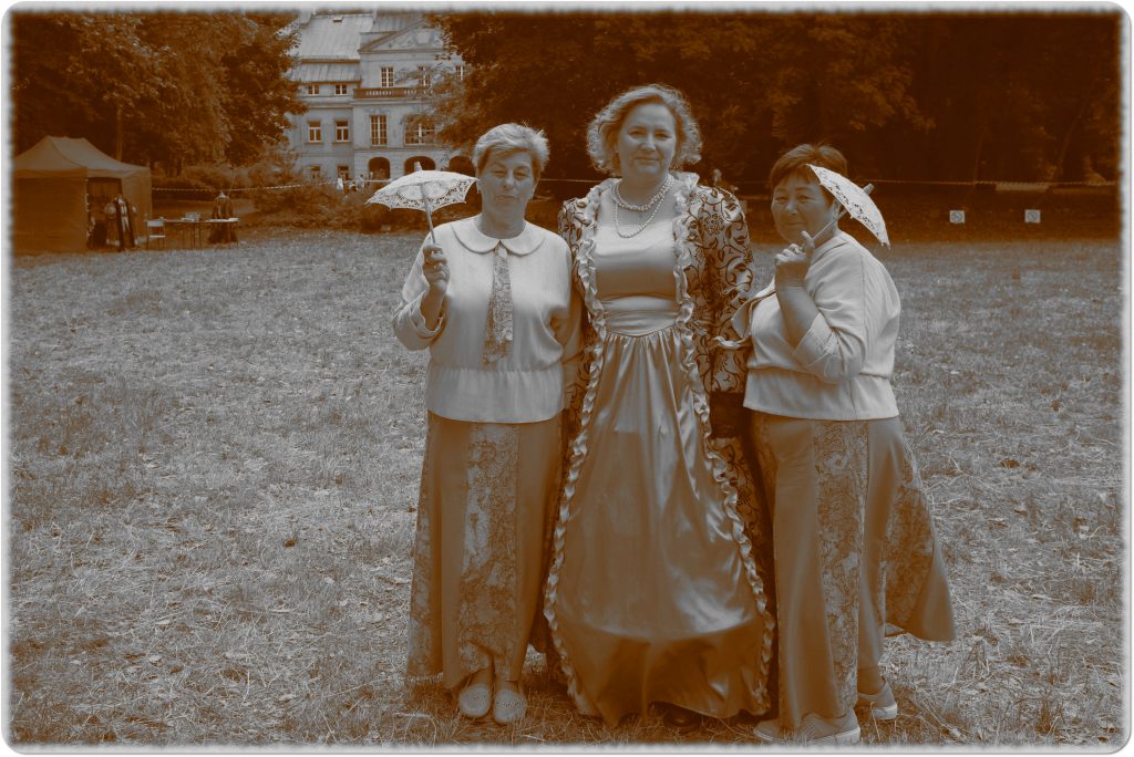 Sepia photo: three smiling women; two holding small umbrellas, the third in a ball gown; behind them, a meadow and a palace