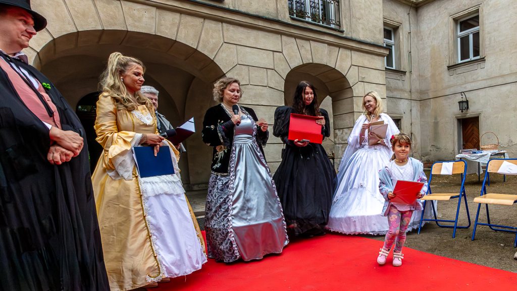 Seven people in historical costumes standing in front of the palace; a girl walks away holding puzzle pieces