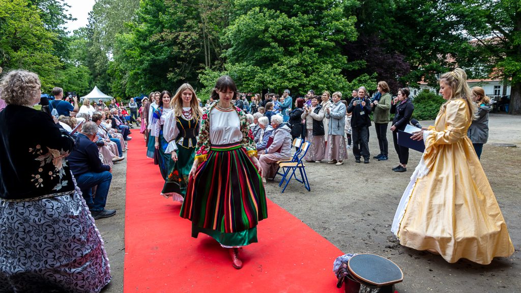 pokaz mody, modeli w strojach balowych idą po czerwonym dywanie wokół nich zebrani widzowie, w tle park
