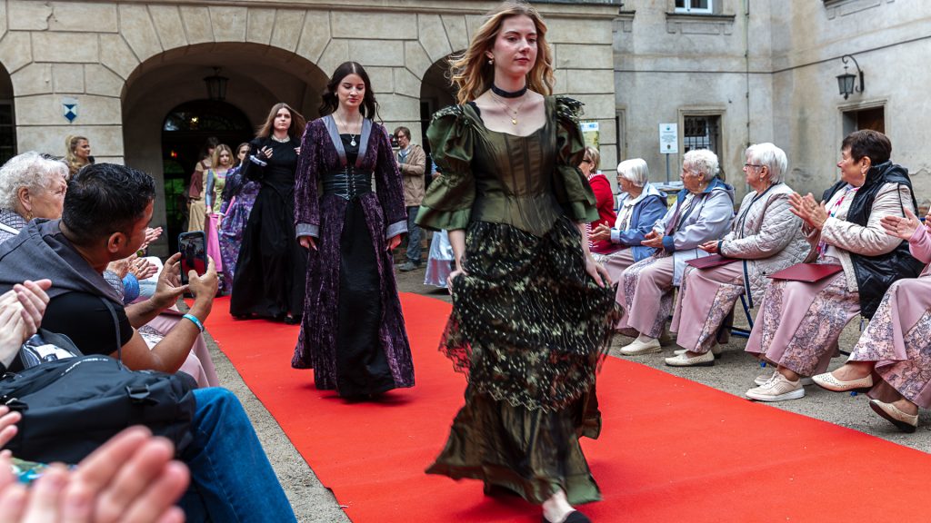 Fashion show: models in ball gowns walking on a red carpet, surrounded by spectators; park in the background