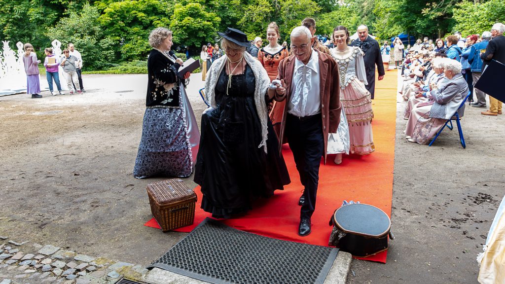 Fashion show: models in historical costumes walking in pairs on a red carpet, surrounded by spectators; park in the background