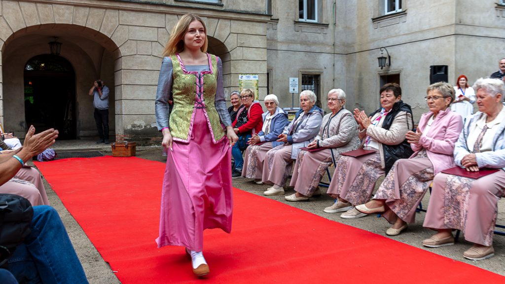 21.	Fashion show: models in ball gowns walking on a red carpet, surrounded by spectators; park in the background