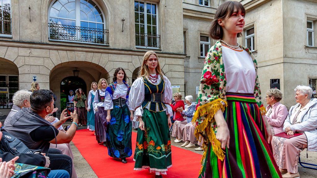 Fashion show: models in folk costumes walking on a red carpet, surrounded by spectators; park in the background