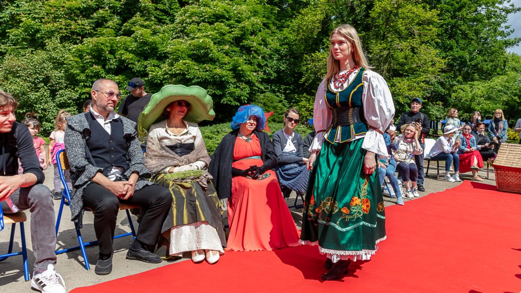 Fashion show: a model in a folk costume walking on a red carpet; in the background, a couple watching the show