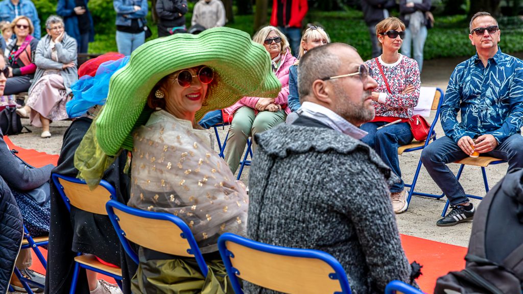 In the foreground, a fancifully dressed couple is seated; in the background, others sit on chairs by the red carpet