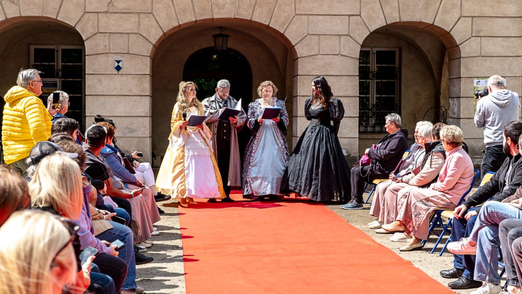 Four people in front of the palace, reading from sheets of paper; audience seated around them