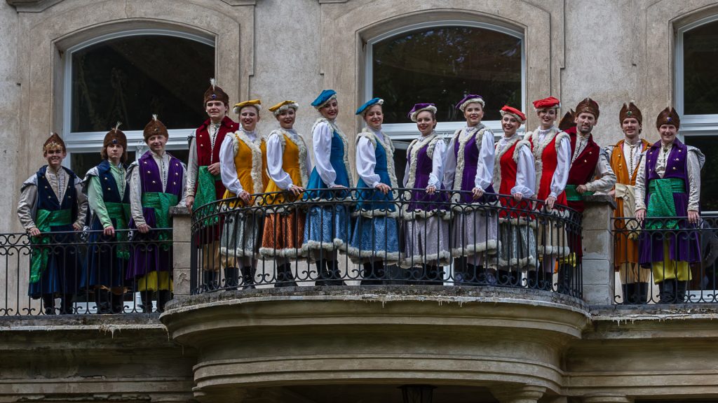 A group of men and women in festive, colorful historical costumes posing in a row on a balcony