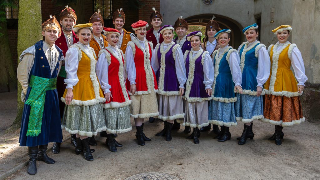 A group of men and women in festive, colorful historical costumes posing in a row in front of the palace