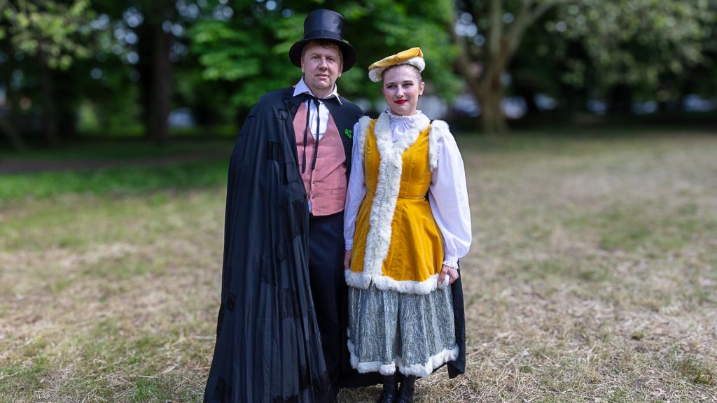 A man in a top hat and long cape posing for a photo with a woman in a folk costume in front of the palace