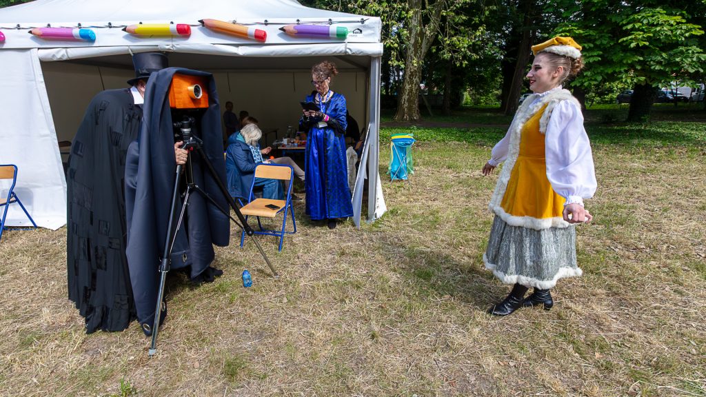 A girl in a folk costume posing for a photo taken with a vintage camera; in the background, a woman with a phone