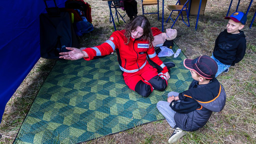 A woman in a red outfit labeled “nurse” showing boys how to administer first aid