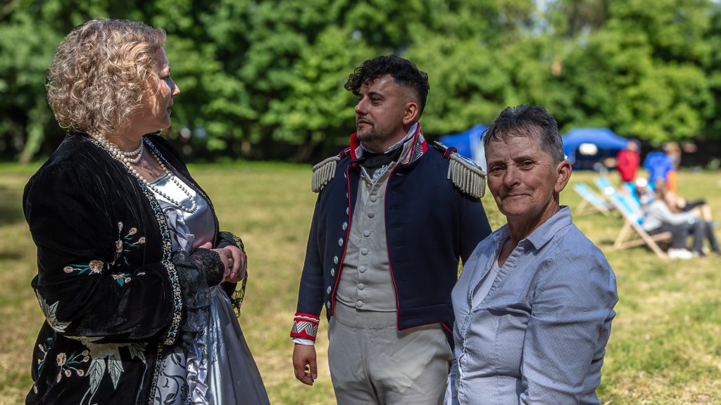 Three people talking in a park setting, two in historical costumes