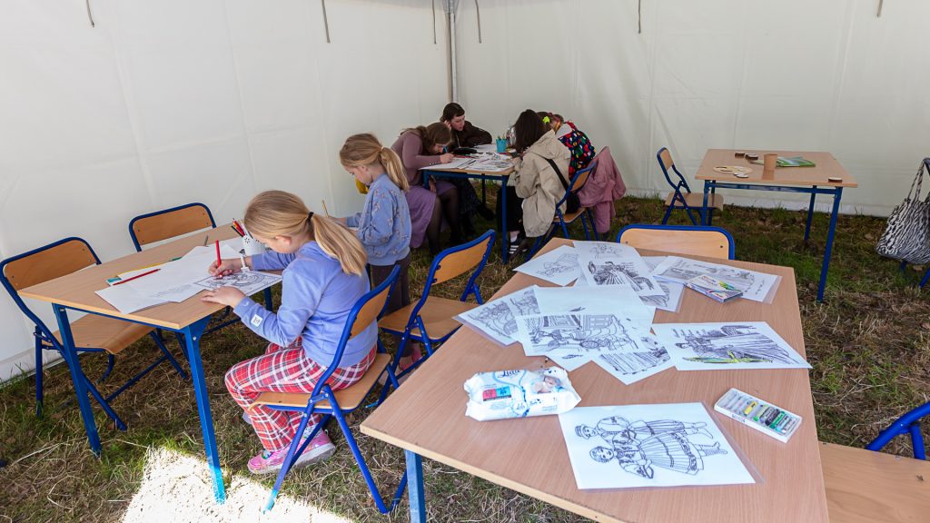 Inside a tent, children sit in groups at tables with art materials