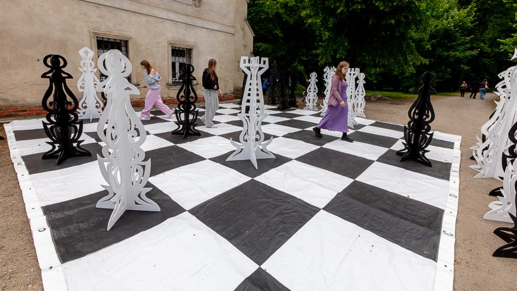 In front of the palace, a giant chess board where girls are moving pieces