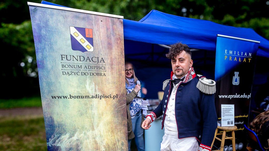 A man in a general’s uniform standing in front of a roll-up banner reading “Bonum Adipisci Foundation – Strive for Good”