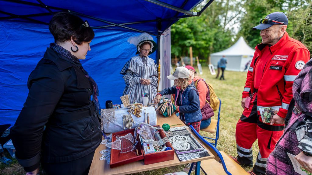 Under a tent, people stand around a table with sewing items; one woman is in historical costume