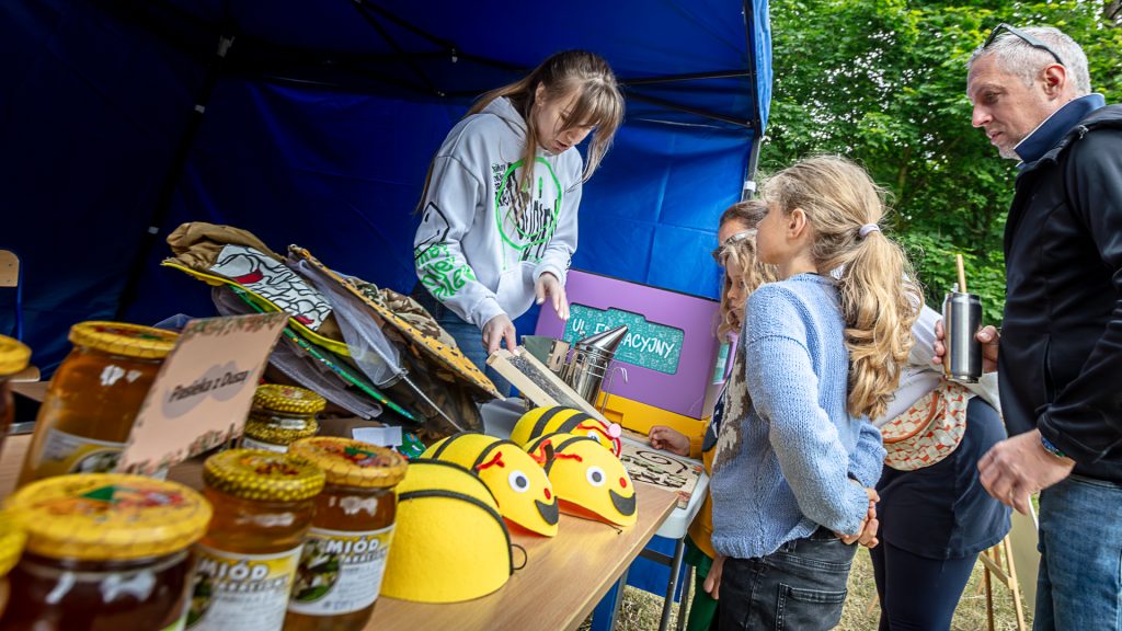 Under a tent, people stand at a table; one person is showing beekeeping-related items