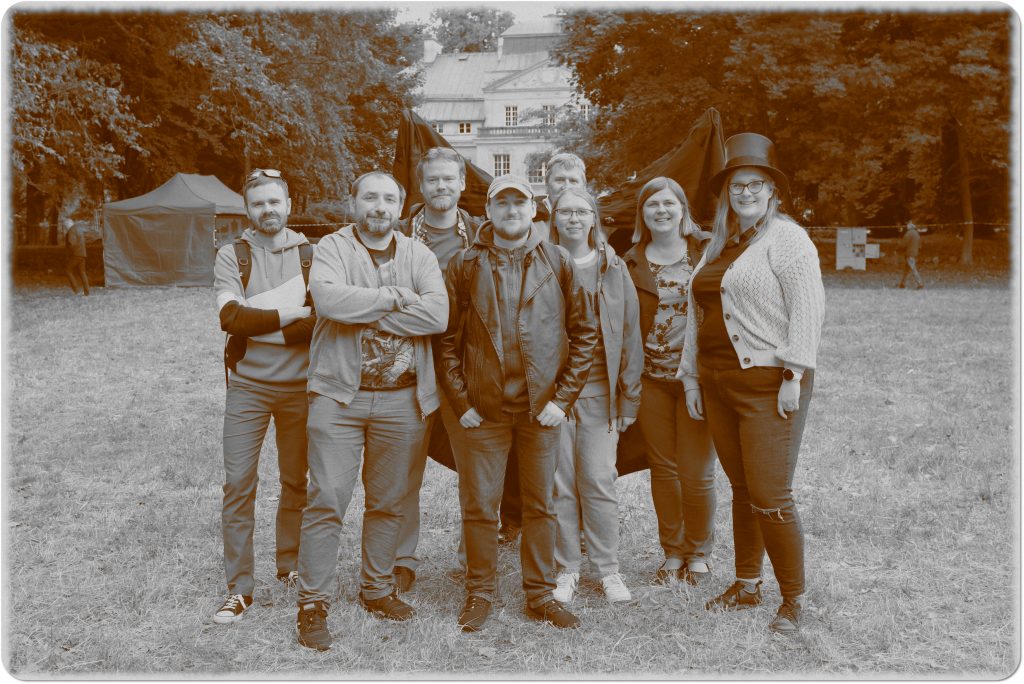 Sepia photo: a group of 8 people, women and men; one of them spreads a large cape behind the group; behind them, a meadow and a palace