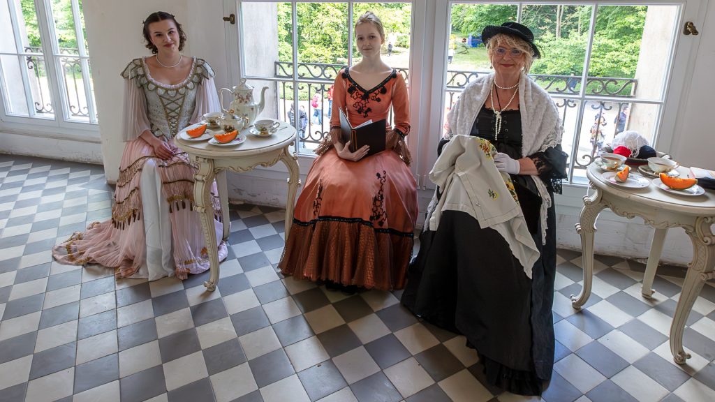 Inside the palace, three women in historical costumes sit in front of a window with a park view