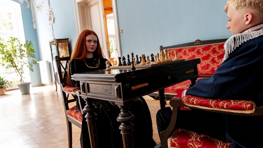 Inside the palace, a boy and a girl in historical costumes playing chess