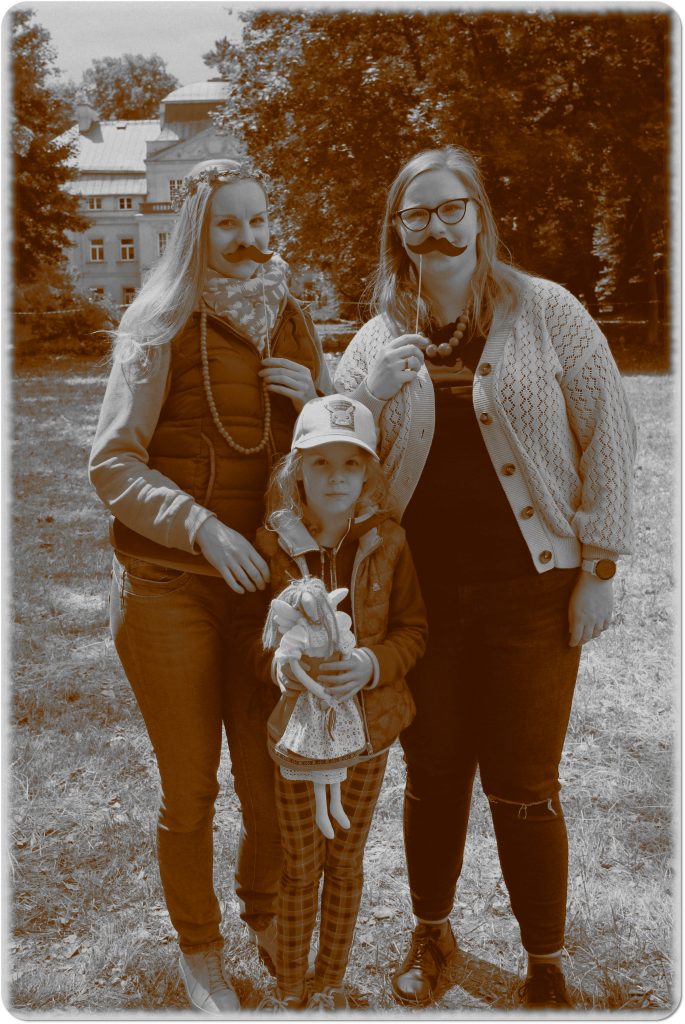 Sepia photo: two smiling women with a child in the middle; behind them, a meadow and a palace