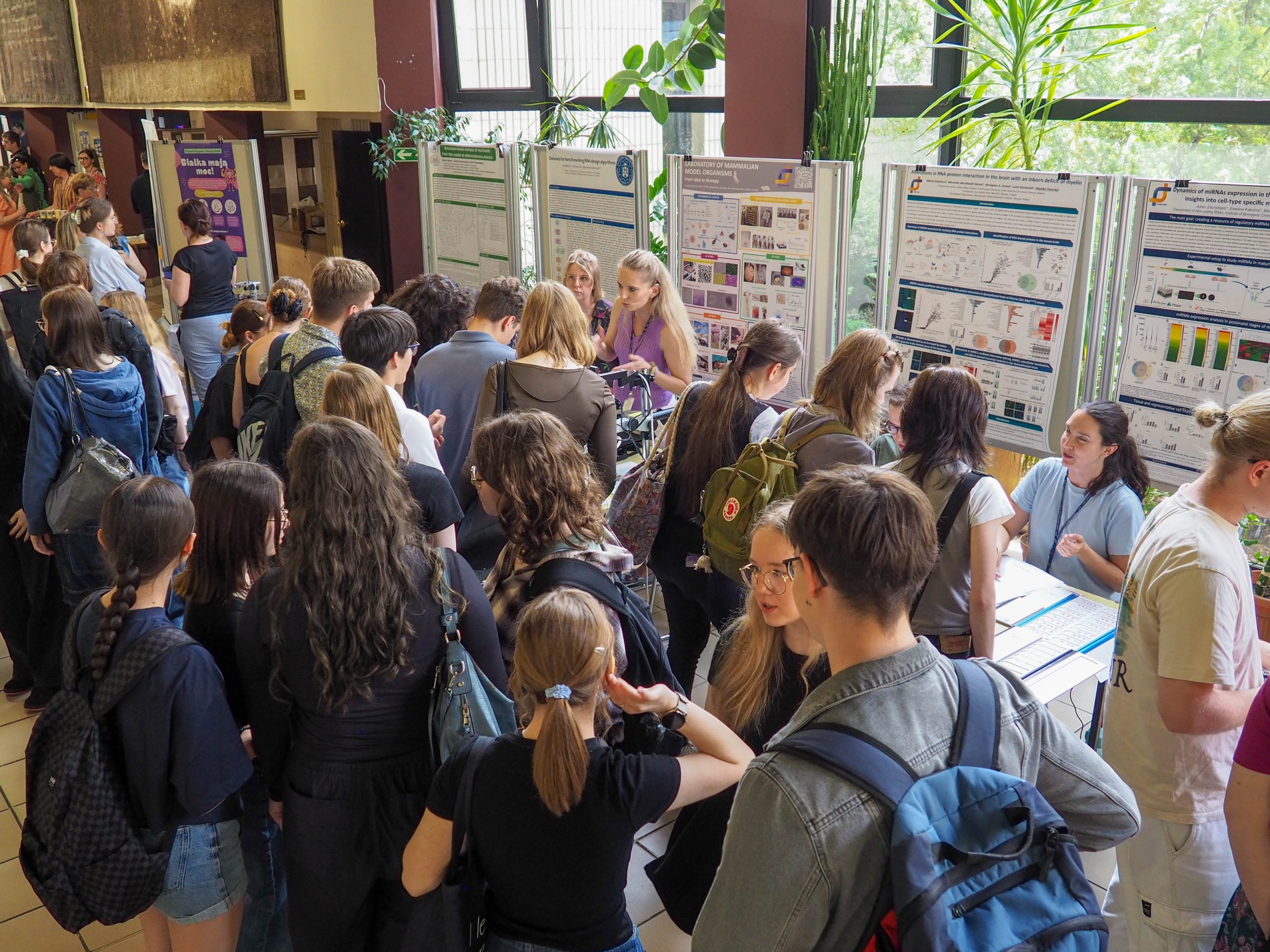 Tłum młodych osób w holu podczas Drzwi Otwartych w ICHB PAN, w tle postery z publikacji naukowych.     Crowd of young people in the lobby during the Open Doors at IBCH PAS, with posters from scientific publications in the background.