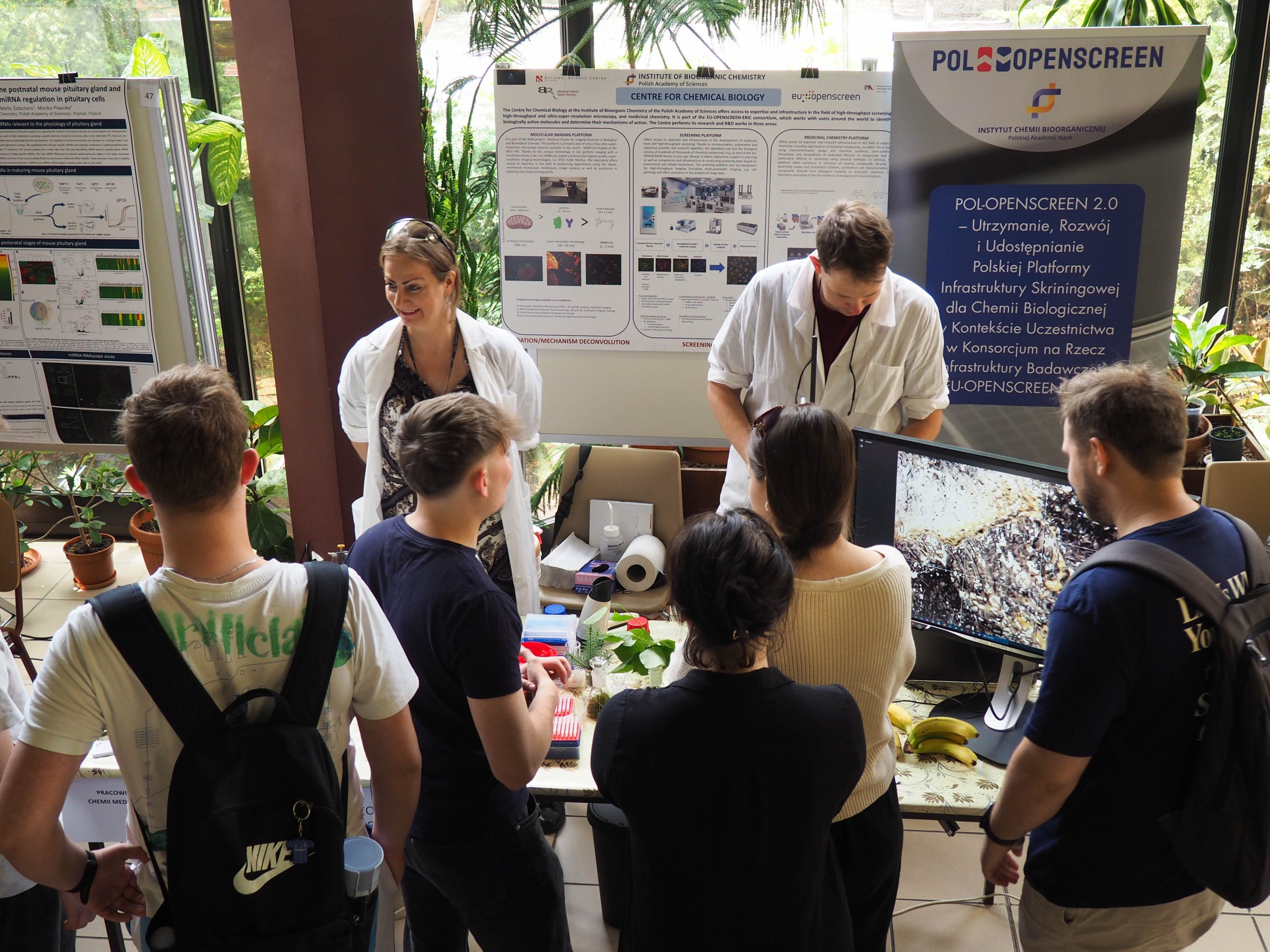 Grupa młodych osób słuchająca i oglądająca z zaciekawieniem stoisko Pracowni Chemii Medycznej.       A group of young people listening and watching the Medicinal Chemistry Laboratory stand with interest.