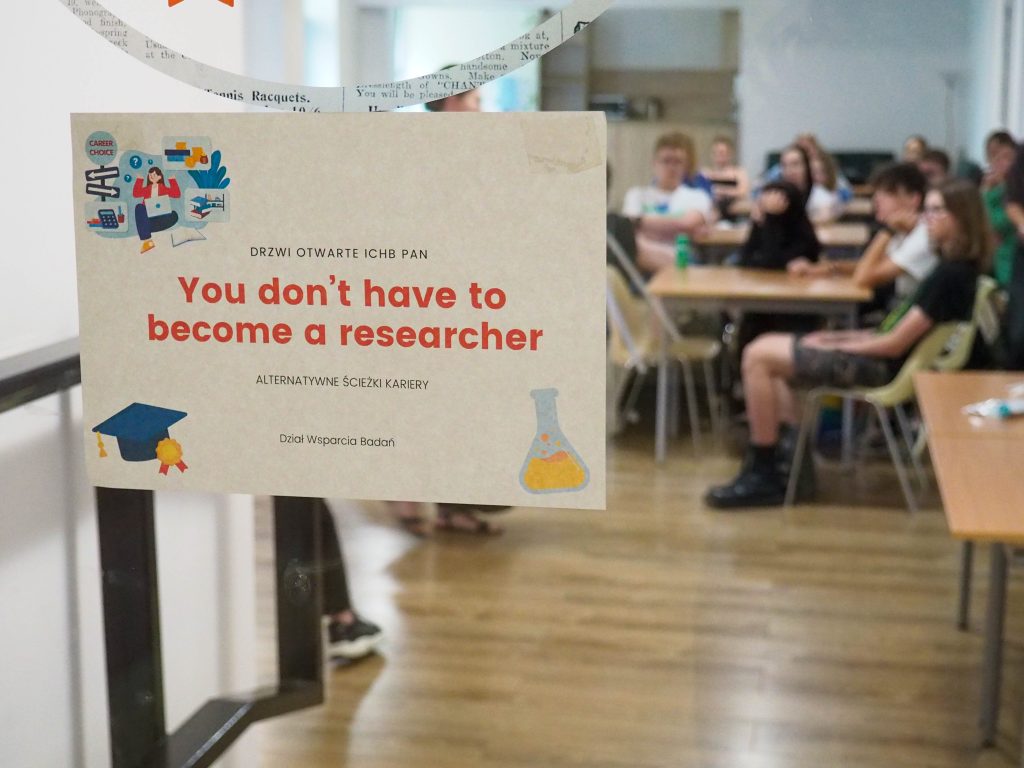 Kartka na drzwiach z napisem You don`t have to become a researcher, w tle grupa osób słuchających prezentacji