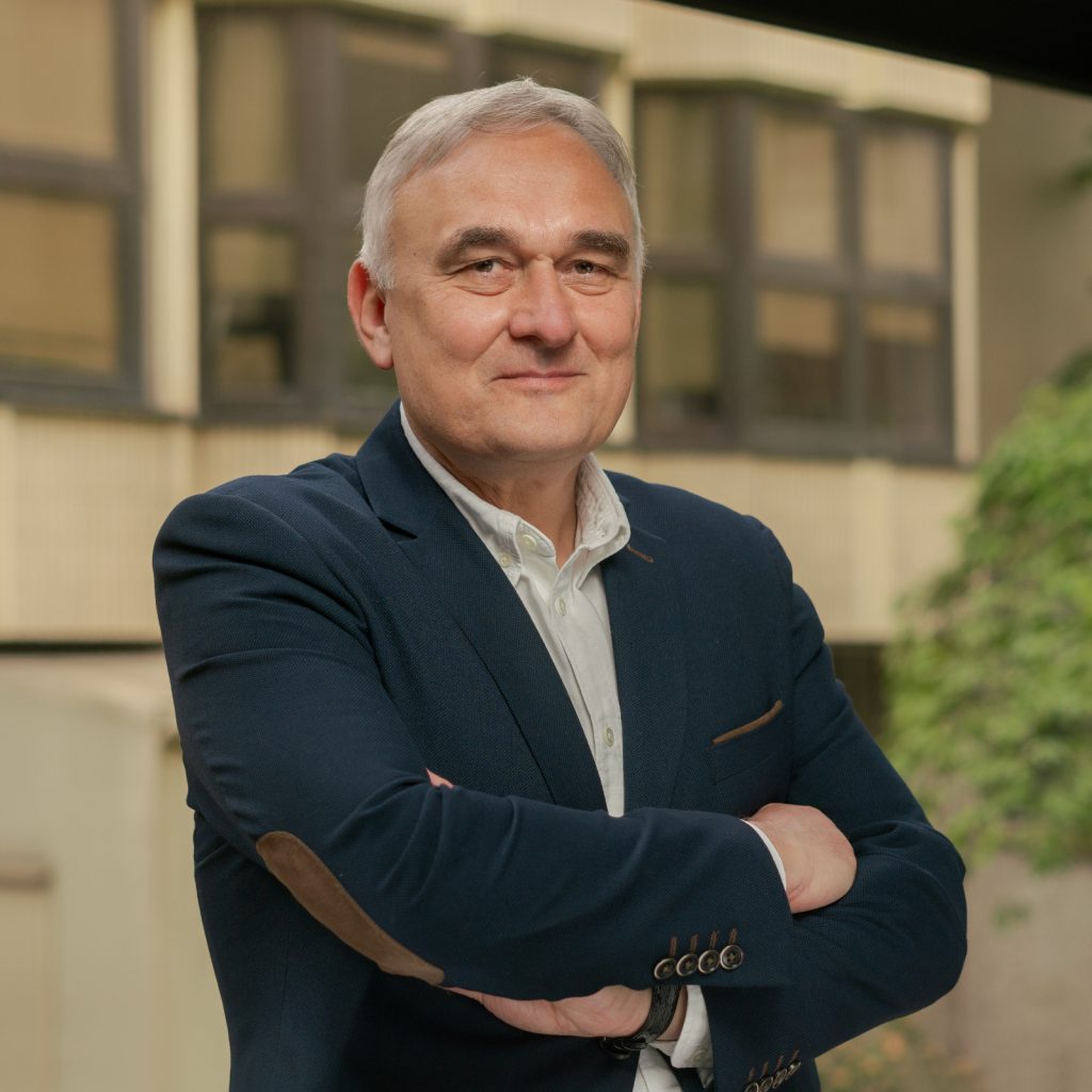 Zdjęcie portretowe prof. Marka Figlerowicza. Mężczyzna w granatowej marynarce z rękoma założonymi na siebie i w białej koszuli, uśmiechnięty. W tle budynek Instytutu Chemii Bioorganicznej PAN.
