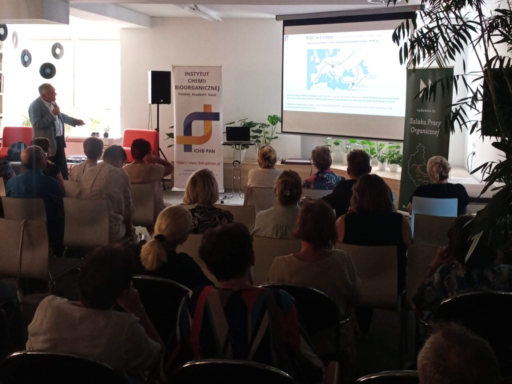 A group of people attentively listens to prof. Marek Figlerowicz, who is delivering a lecture titled "What do we inherit from our ancestors?"