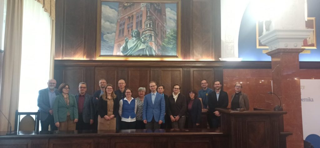 Grupa pozujących do zdjęcia osób przy mównicy w sali uniwersyteckiej, w tle obraz Mikołaja Kopernika