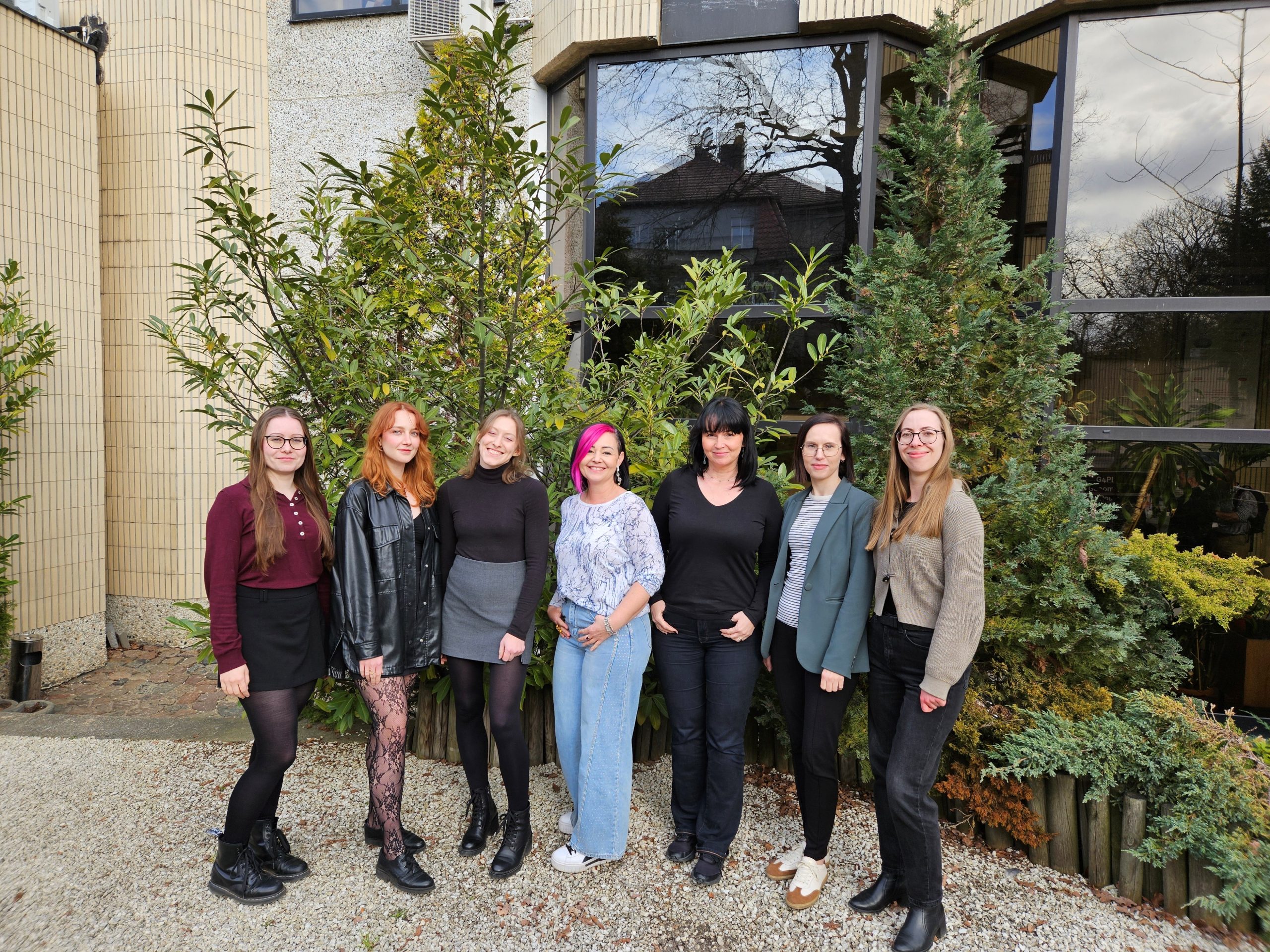 Seven members of the Laboratory of Model Invertebrate Organisms stand next to each other outside the building of the Institute of Bioorganic Chemistry of the Polish Academy of Sciences in Poznań. The group poses on a gravel path in front of a modern building with large windows, surrounded by green shrubs and trees. The women are dressed in casual, semi-formal clothing and are looking toward the camera.