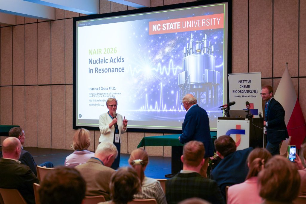 Prof. Zofia Gdaniec speaking into a microphone to the conference participants. In the background, one of the presentations shown during the conference is visible