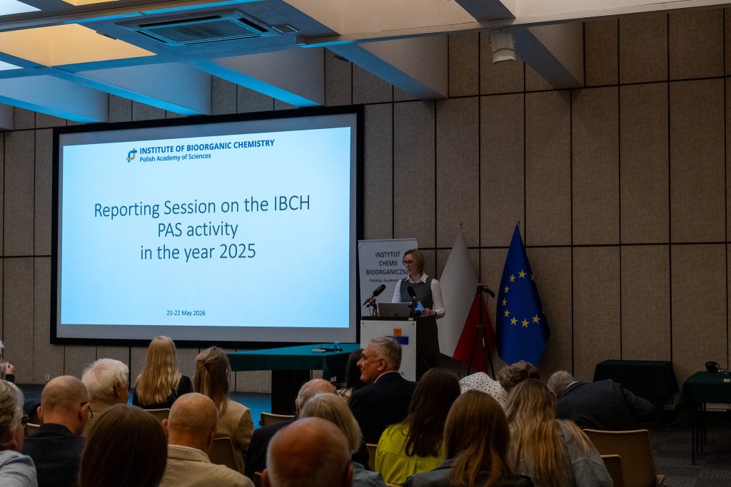 Dyrektor Instytutu przemawia zza mównicy z logo ICHB PAN przed dużym ekranem z napisem zapowiadającym sesję sprawozdawczą za rok 2025. W tle widoczne są flagi Polski i Unii Europejskiej oraz zgromadzona publiczność.