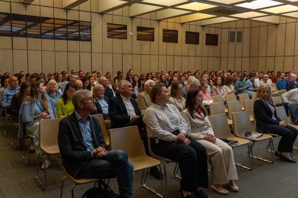 : Szerokie ujęcie dużej sali konferencyjnej wypełnionej siedzącą publicznością. Uczestnicy w rzędach krzeseł patrzą w stronę sceny.