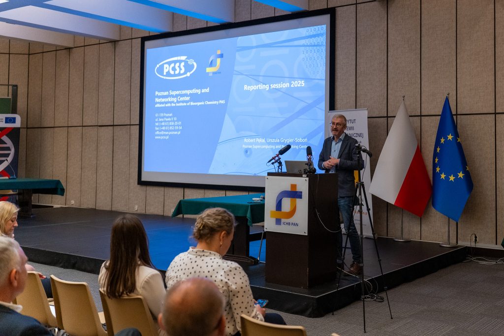 Mężczyzna w marynarce przemawia zza mównicy na tle ekranu z prezentacją Poznańskiego Centrum Superkomputerowo-Sieciowego (PCSS). Obok sceny stoją flagi Polski i Unii Europejskiej.
