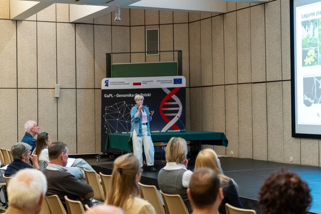 A woman in a denim shirt speaks on stage with a microphone, standing in front of a backdrop that reads "G4PL - Genomika dla Polski". A portion of a scientific presentation screen is visible on the right.