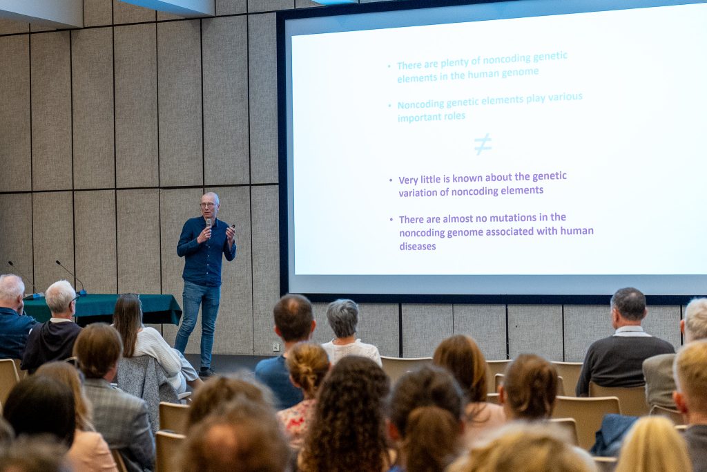 : A man in a dark blue shirt gestures while speaking on stage with a microphone. Next to him, a large screen displays a presentation about the human genome.