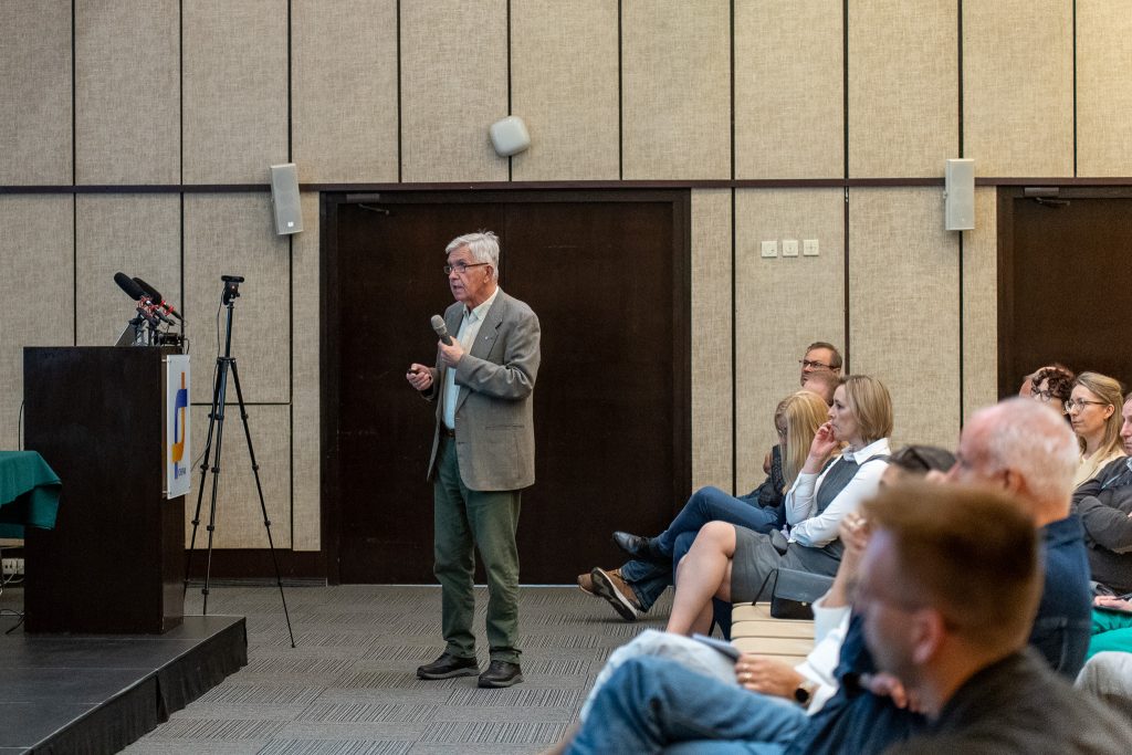 : A man in a gray blazer stands in the middle of the hall with a microphone, speaking to the seated attendees. A portion of the institute's lectern is visible on the left.