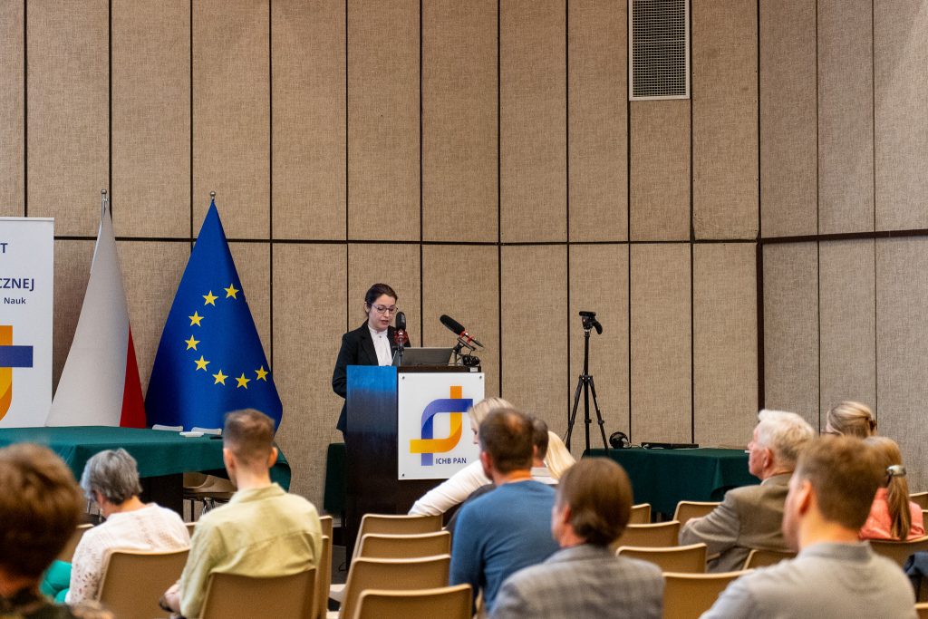 Kobieta w ciemnej marynarce stoi za mównicą z logo ICHB PAN i przemawia. W tle po lewej stronie widać flagi Polski i Unii Europejskiej.