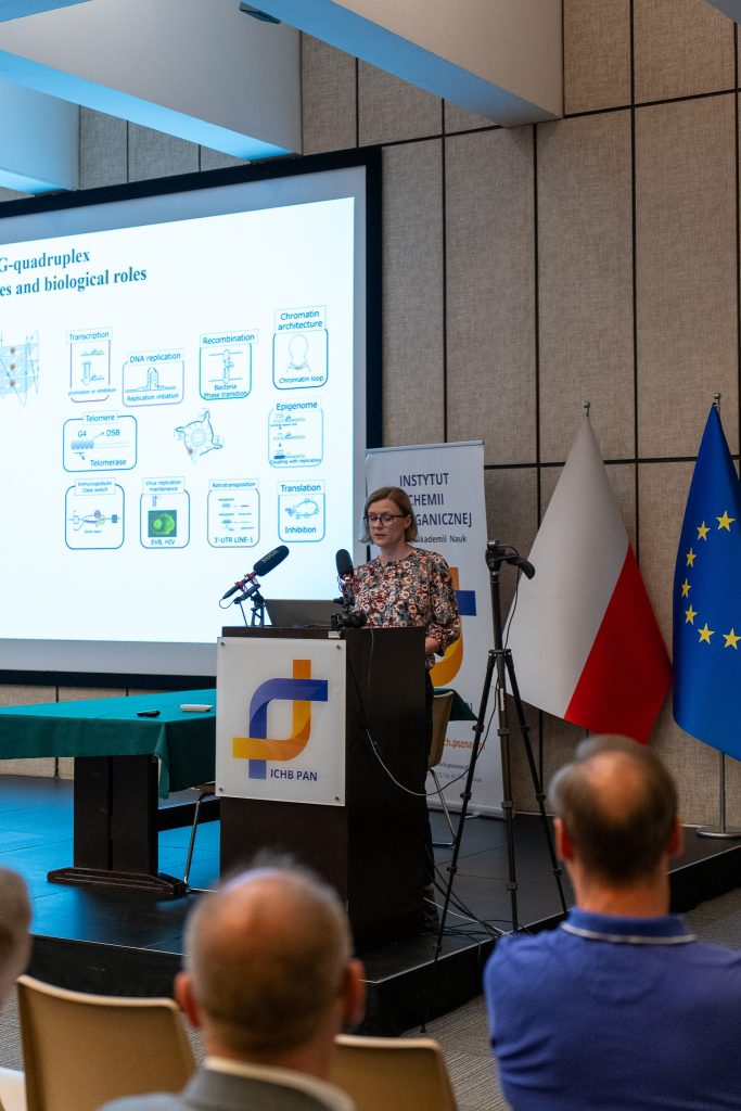 Kobieta we wzorzystej bluzce przemawia zza mównicy, za którą wyświetlany jest schemat struktur biologicznych. Obok sceny stoją flagi Polski oraz Unii Europejskiej.