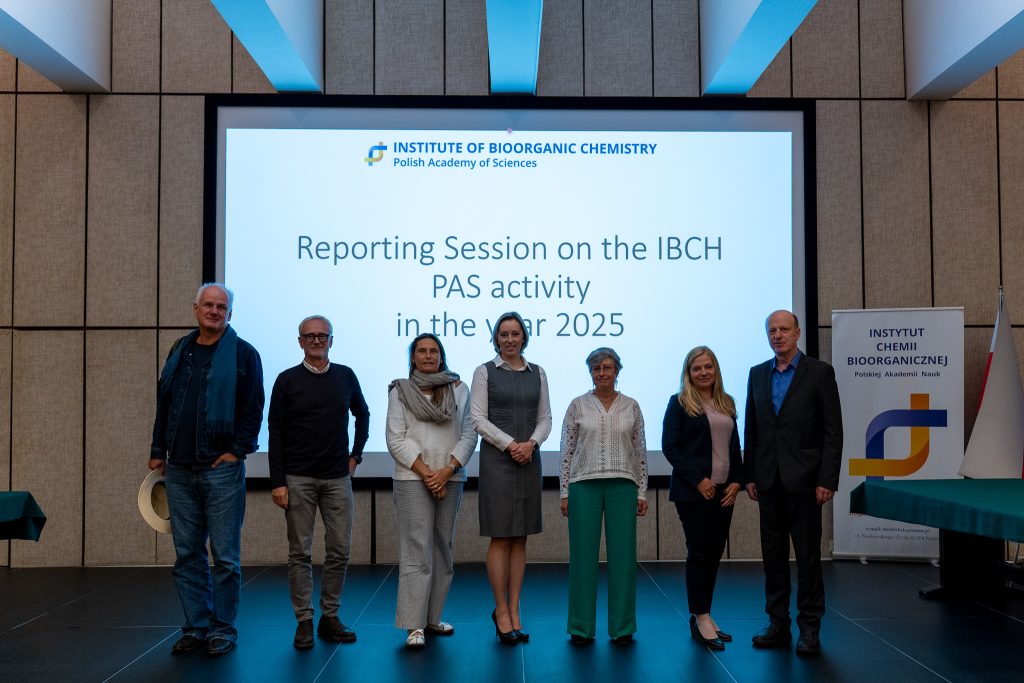 Siedmioro uczestników stoi w rzędzie na scenie do pamiątkowego zdjęcia na tle ekranu z napisem „Reporting Session on the IBCH PAS activity in the year 2025”. Po prawej stronie widoczny jest pionowy baner informacyjny instytutu.