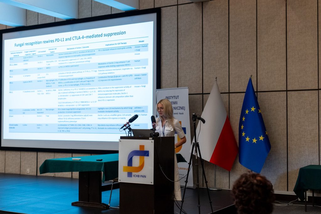 A woman in light dress speaks from behind a lectern with the ICHB PAN logo. Behind her, a large screen displays a scientific presentation.