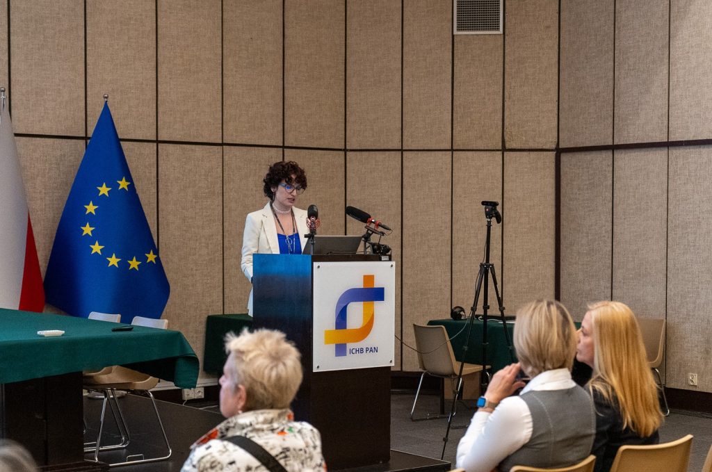 A woman with dark hair, wearing a blazer, stands on stage behind the lectern. Next to her, flags are visible.