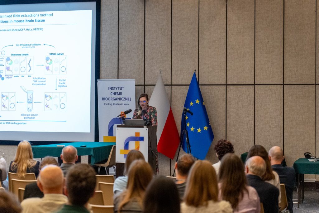 A woman in dark deress speaks from behind the official institute lectern. A slide summarizing projects and scientific publications is displayed on the screen in the background.