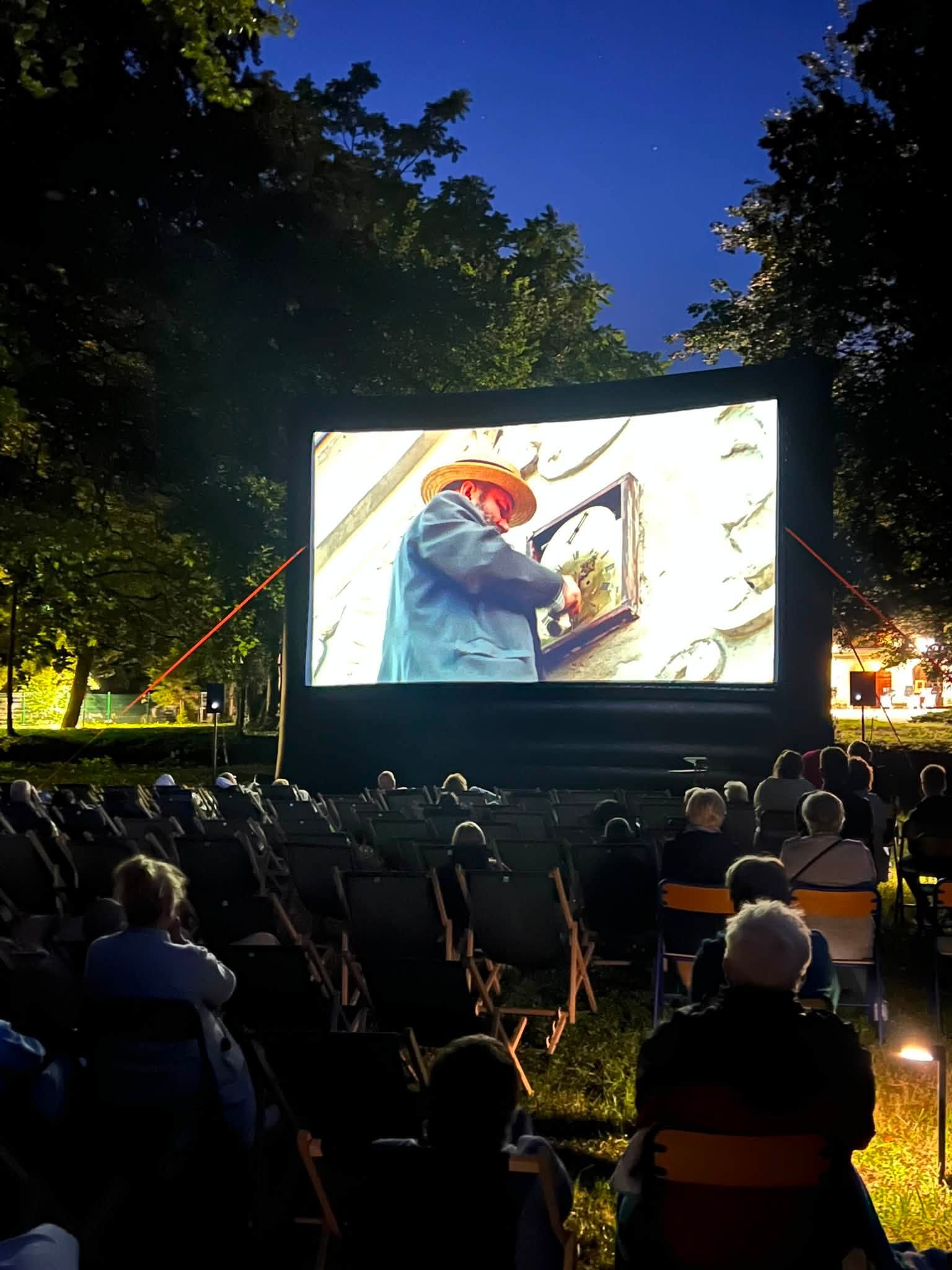 Wieczorne kino plenerowe w parku. Na dużym ekranie wyświetlany jest film, a przed nim siedzą widzowie na leżakach. Wokół znajdują się oświetlone drzewa, a w tle widać ciemne, wieczorne niebo.