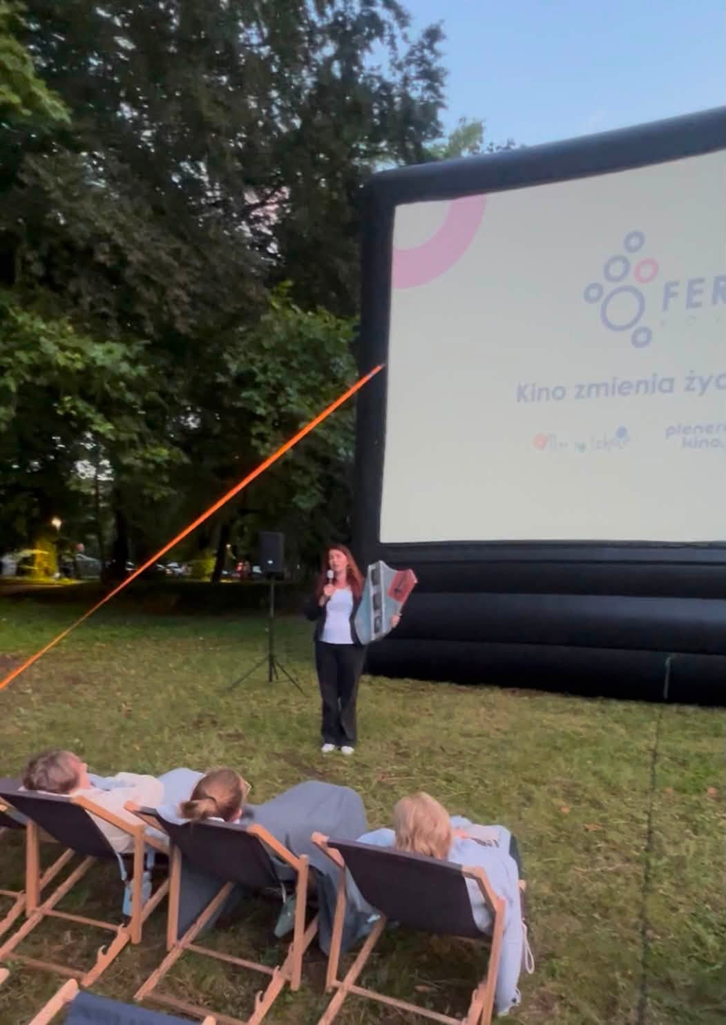 Kobieta stojąca przed ekranem podczas kina plenerowego, trzymająca w ręku herb. W tle duży ekran projekcyjny, parkowe drzewa i widzowie siedzący na leżakach