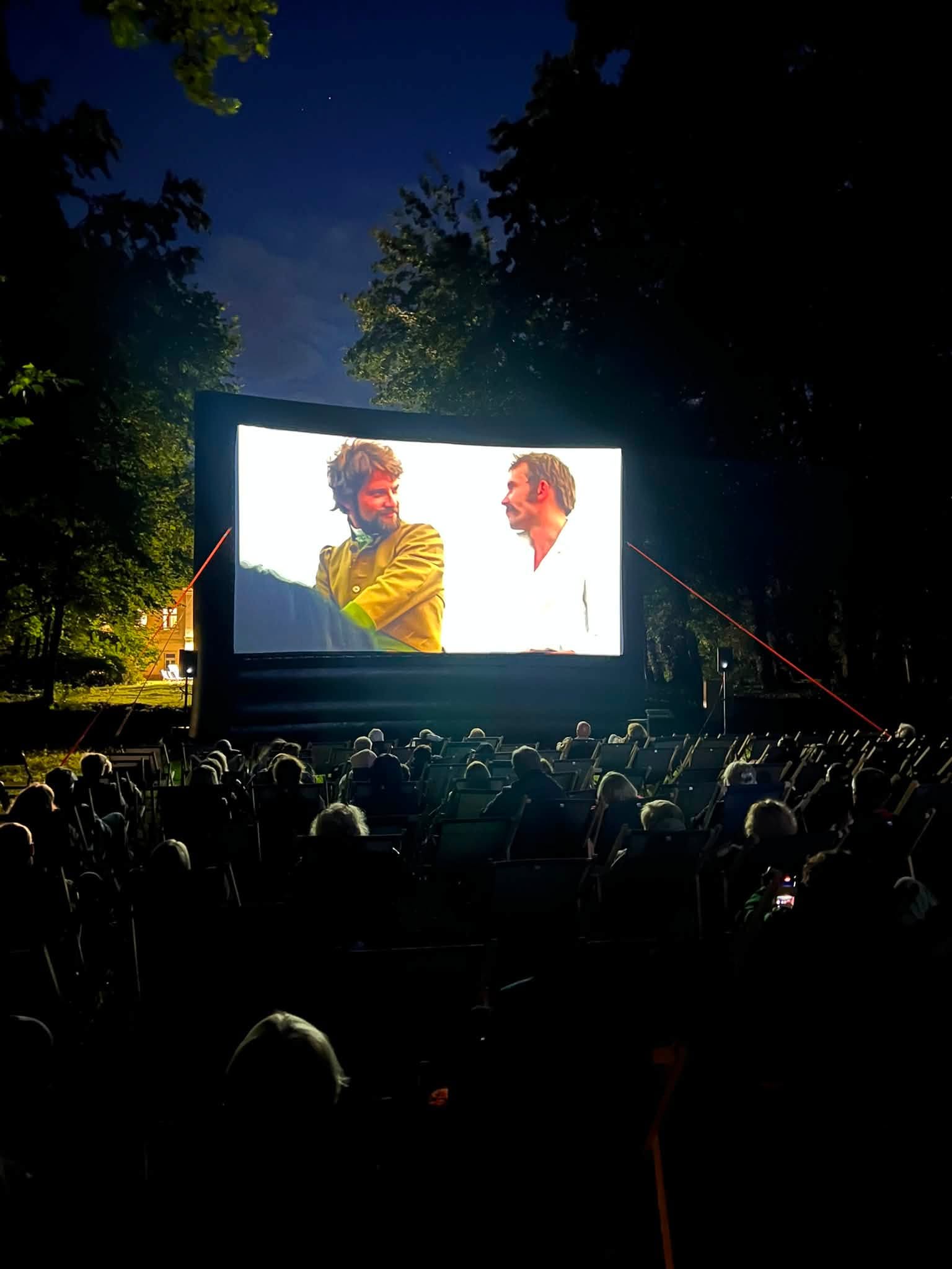 Wieczorne kino plenerowe w parku. Na dużym ekranie wyświetlany jest film, a przed nim siedzą widzowie na leżakach. Wokół znajdują się oświetlone drzewa, a w tle widać ciemne, wieczorne niebo.